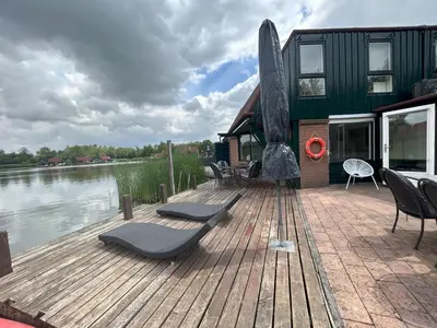 a wooden deck with chairs and a house on the water at Lakeside Cottage De Rijd in Nieuwe-Niedorp