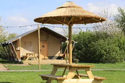 a picnic table with a straw umbrella in front of a hut at Safari Lodges in Nieuwe Wetering