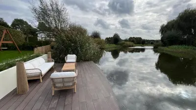 a wooden deck with a couch and chairs next to a river at Villa Liferla - Wellness met sauna en jacuzzi in Zeewolde