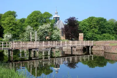 a bridge over a river with a church in the background at De Slotruine in Egmond aan den Hoef