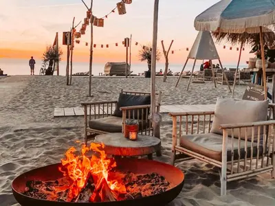 a fire pit on a beach with chairs and an umbrella at La Siesta Noordwijk Zee in Noordwijk